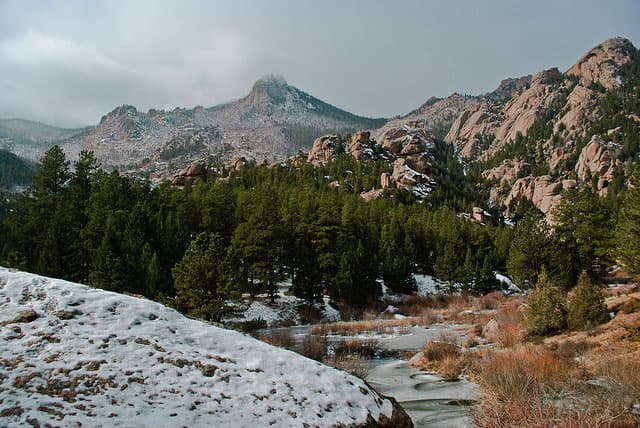A small creek is frozen over with ice near Bailey, Colorado. A thick stand of pine trees are on the far bank with rocky peaks rising up behind them.