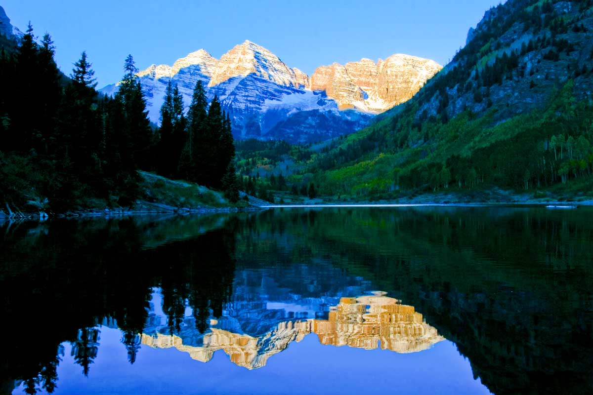 The Maroon Bells peaks are painted yellow and blue by the fading sunlight. Their image is reflected in Maroon Lake near Aspen and Snowmass.