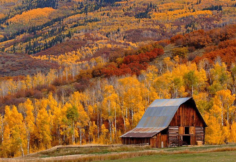 A famous red barn sits in front of a mountain covered with trees in fall foliage — they are rich shades of crimson, orange and gold