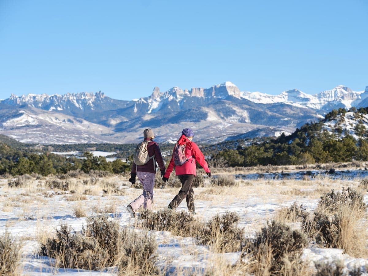 Two people walk along a snowy trail under blue skies in Ridgway with jagged peaks in the distance