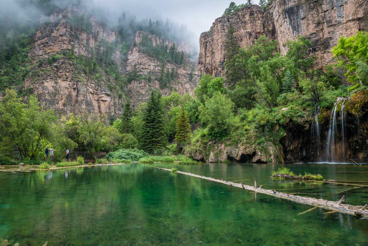 A fallen tree lies across a serene lake reflecting the verdant green trees surrounding it near Glenwood Springs. A gentle stream of water cascades down from a rock wall and feeds the lake. Overhead, fog hangs around towering peaks.