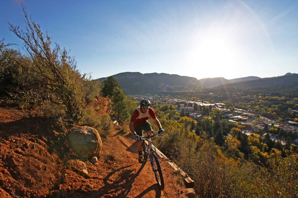 Beneath a setting sun, a mountain biker rides a narrow trail. Downtown Durango can be seen below.