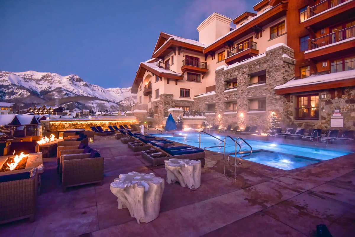 A large steaming spa and pool and hot tub filled with blue water sits among fire pits, blue-cushioned seats and other stools and decorative items for the outside terrace. Behind the pool is the Madeline Hotel & Residences of Telluride, the buildings adorned with stone and dark wood.