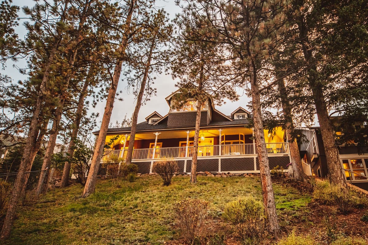 As the day ends, the lights of the Outlook Lodge wraparound porch turn on. Pine trees stand tall around the lodge.