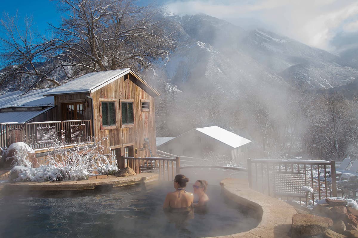 Two bathers soak in the steamy waters of a hot-springs pool at Avalanche Ranch near Redstone, Colorado. In the background, a snowcapped peak rises above the floating steam.