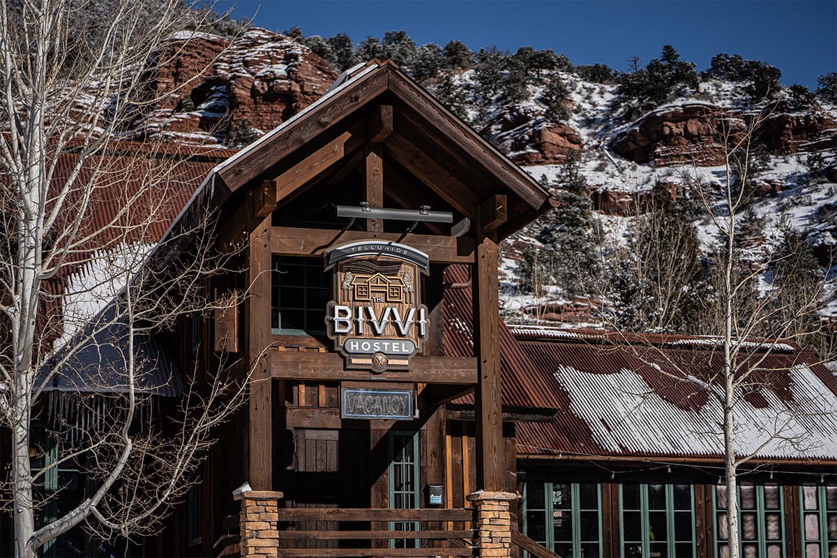 A dark-wood lodge style building is set beneath tall red-rock cliffs dotted with pine trees. The Colorado building and cliffs have a layer of snow on them.