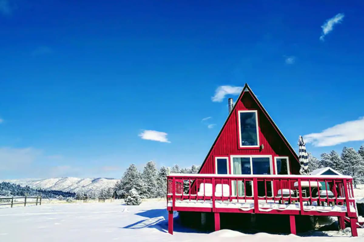 A cherry-red A-frame chalet is covered in a blanket of snow in Colorado. Overhead are bright blue skies dotted with a few clouds.