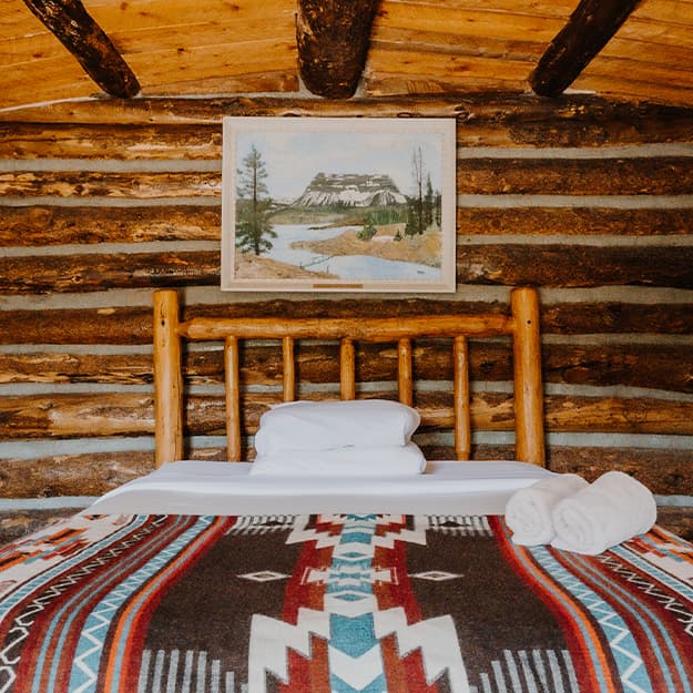 A log-frame bed sits beneath a painting of a local butte at Trappers Lake Lodge. The room is in a log cabin with thick, exposed beams and a wood-panel ceiling.