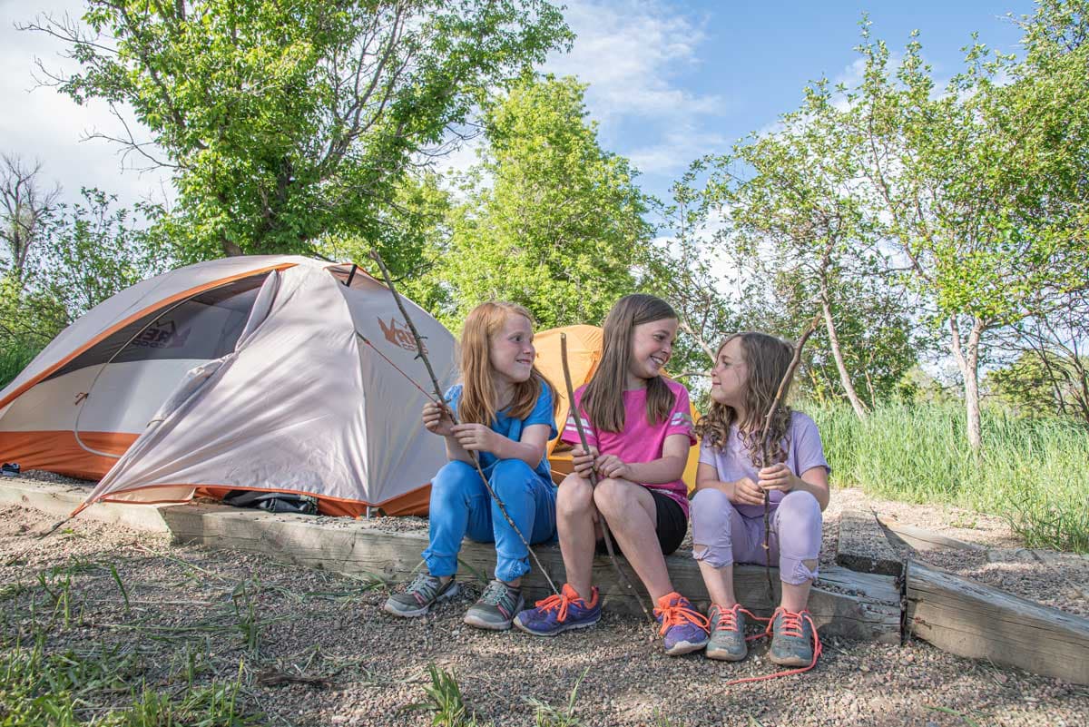 Three girls sit on a log holding sticks near a tent at Yampa River State Park.