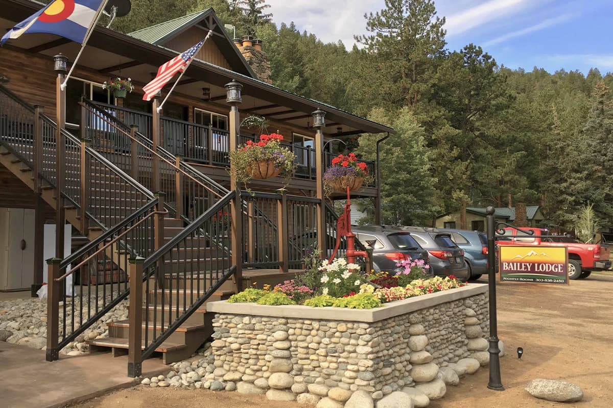 The front of the Bailey Lodge in Colorado looks picturesque with flower pots and a wide porch, along with a blue cloud-streaked skies and thick pine forest in the background.