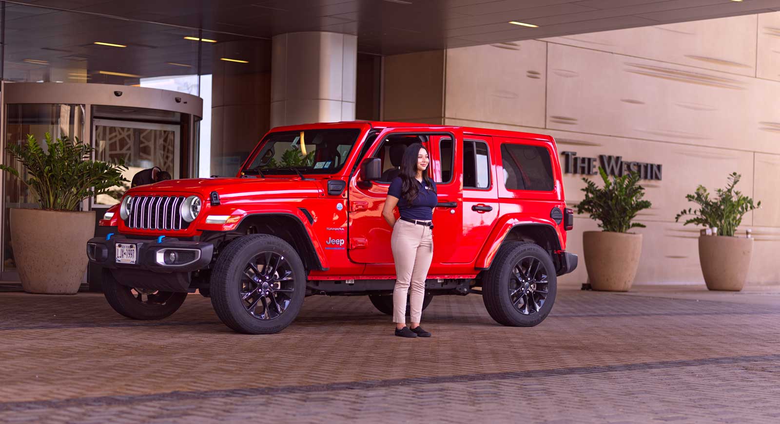 A woman stands in front of a red Jeep at a hotel