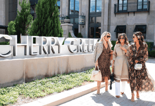 Cherry Creek shopping mall in the background with large "Cherry Creek" sign in the front and three women dressed up talking to each other walking past the sign.