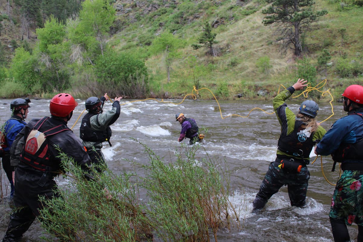 Six students in protective gear and backpacks stand knee-deep in whitewater waves of a river in Colorado. One of them is in the middle of the river while the rest are more on the side of it. Between them, there is a thin yellow rope frozen in the air, being tossed between them as part of the course. In the background is a tall, steep, green hill with scattered small trees and rocks.