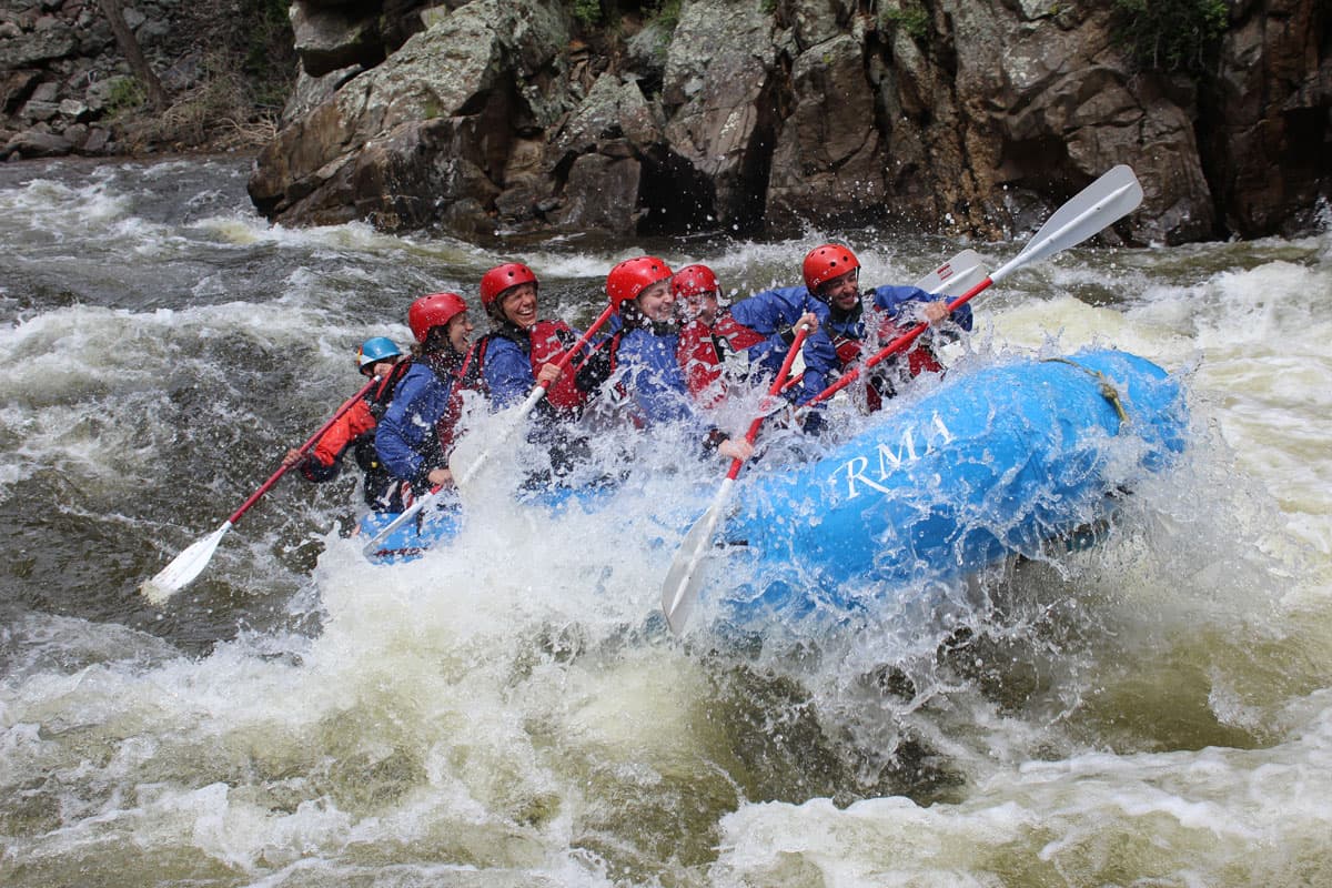 A group of whitewater rafters in red protective gear smiling and laughing on a blue raft. The raft is being tossed by the rushing water, splashing the people's faces. They all have paddles in their hands. Behind them on the raft is the instructor, barely visible, with their paddle in the water. In the background are rocky cliff-like structures beside the water.