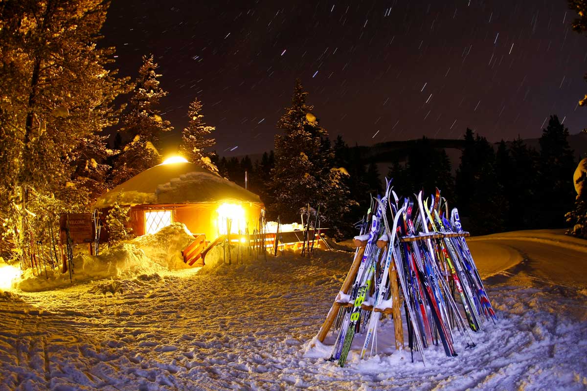 Skis are stacked in the snow in front of a yurt lit up in the background at night