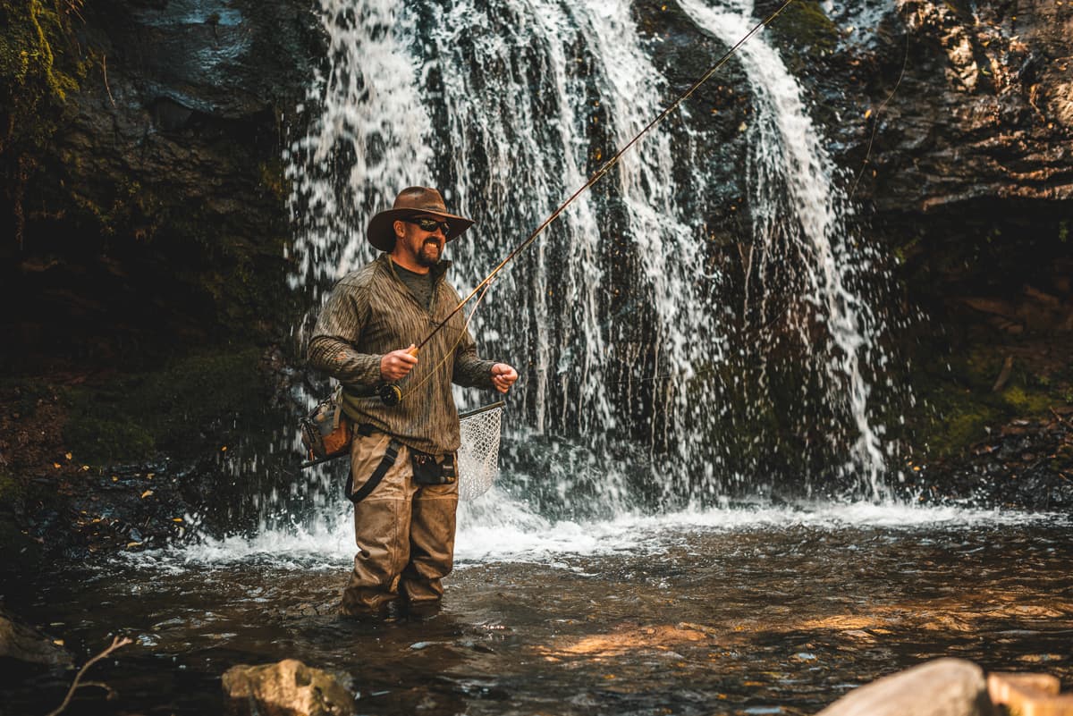 Someone fishes in a river with their handcrafted fly-fishing bamboo rod. They are shin-deep in the water and are smiling with a hat and sunglasses on. In the background is a small waterfall.