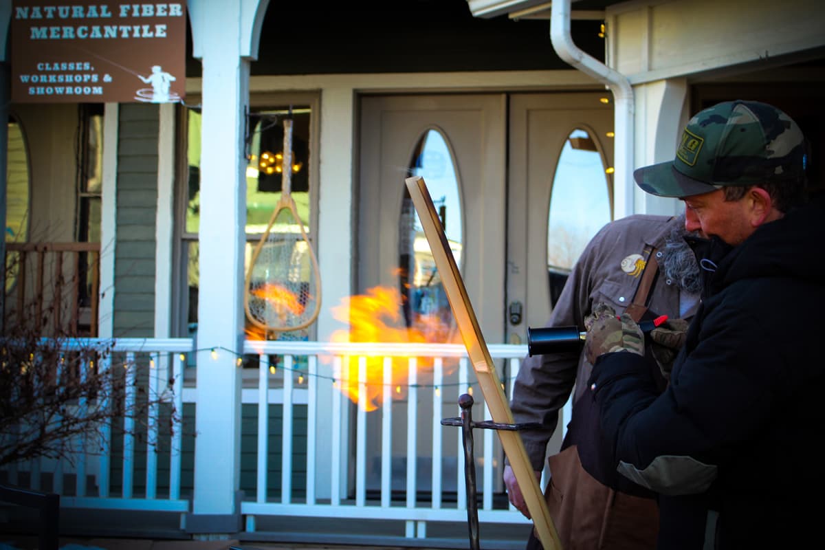 A photo of two people outside the PJ's Fine Bamboo Rods workshop in Pagosa Springs, Colorado. In the foreground are two people using a blowtorch to heat treat bamboo, and the flame is orange and yellow. In the background are the doors with reflective glass windows of the Natural Fiber & Mercantile.