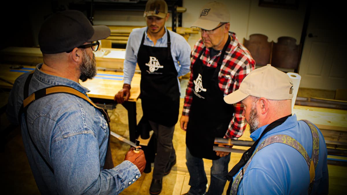 Four people stand in a workshop in PJ's Fine Bamboo Rods in Pagosa Springs. At least one of the people is an instructor, who shows the students what to do.