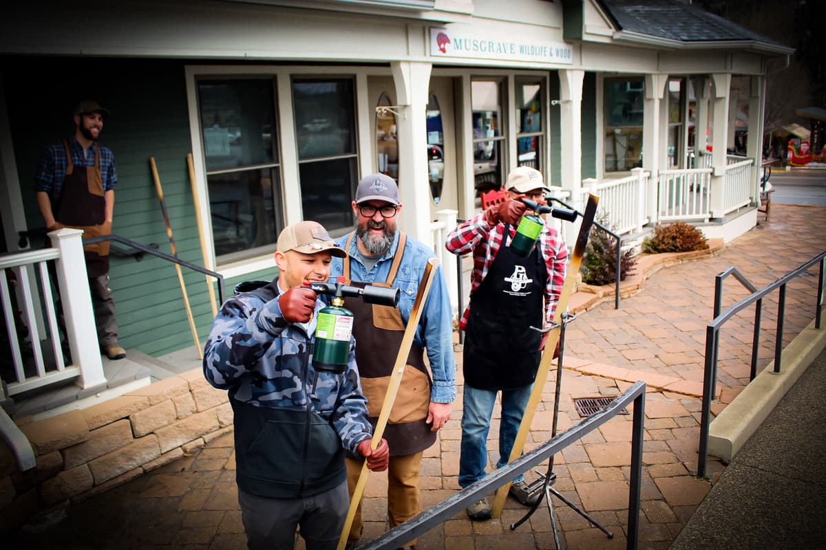 Three people, including the instructor of the class, stand outside in warm clothing outside the PJ's Fine Bamboo Rods workshop in Pagosa Springs. They are all smiling as they heat-treat their bamboo strips. In the background are more storefronts.