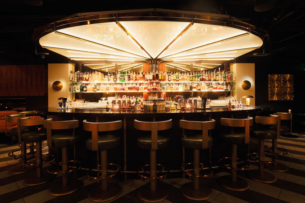 An intricate half-circular bar with bar stools surrounding the marble counter top. The lighting is generally dim but the large circular lighting fixture on the ceiling bathes the fully stocked bar in a warm glow. There are gold-colored decorative wall fixtures surrounding the bar.