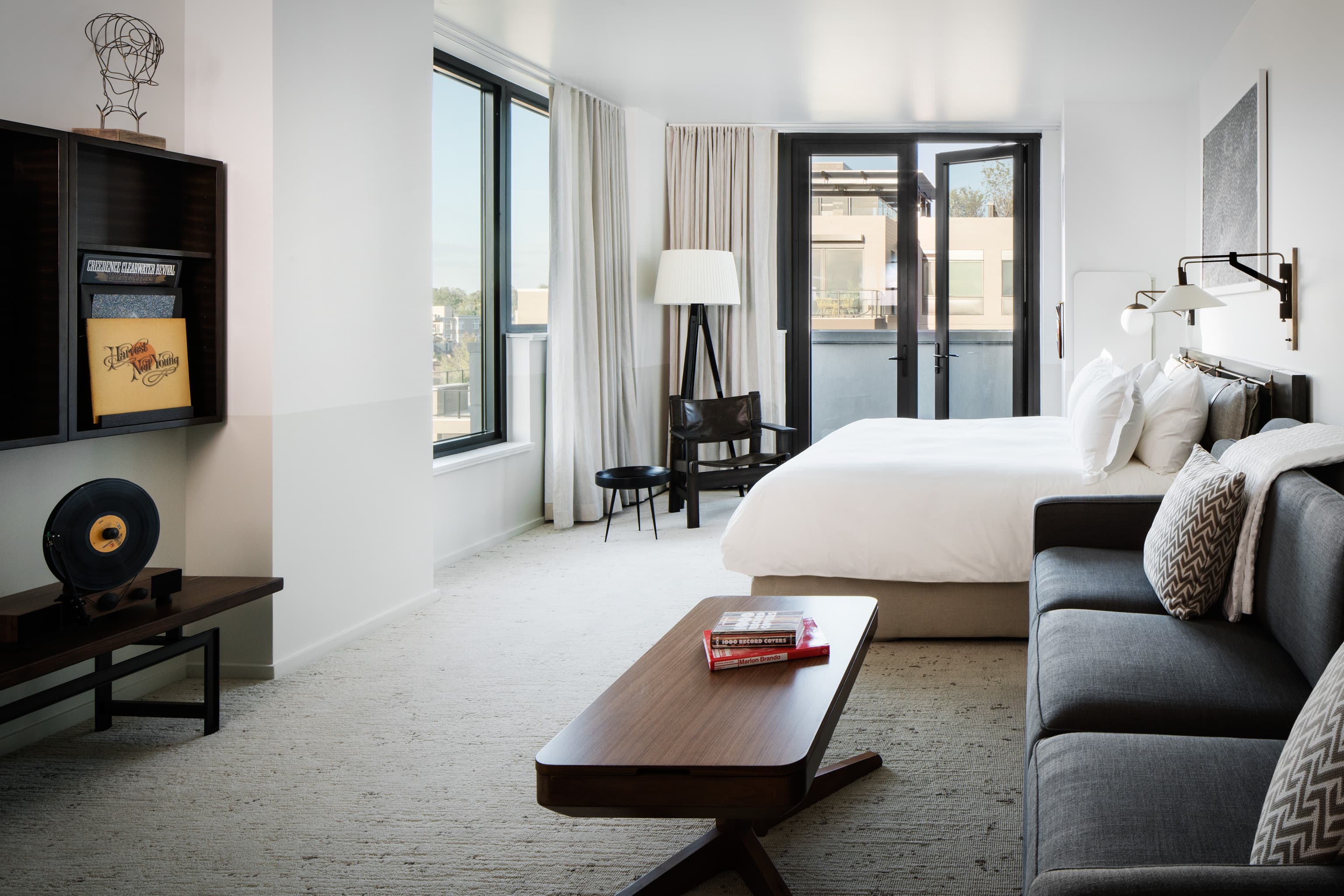 A photo of a clean, crisp room at the Halcyon hotel in Denver. In the front right corner of the photo is a three-seat dark gray couch with decorative pillows in front of a modernly decorated wall shelf. Next to the couch is a full bed dressed in white linens. On the far wall is an open patio door overlooking the city, and the wall next to it has a tall window also looking over the city.