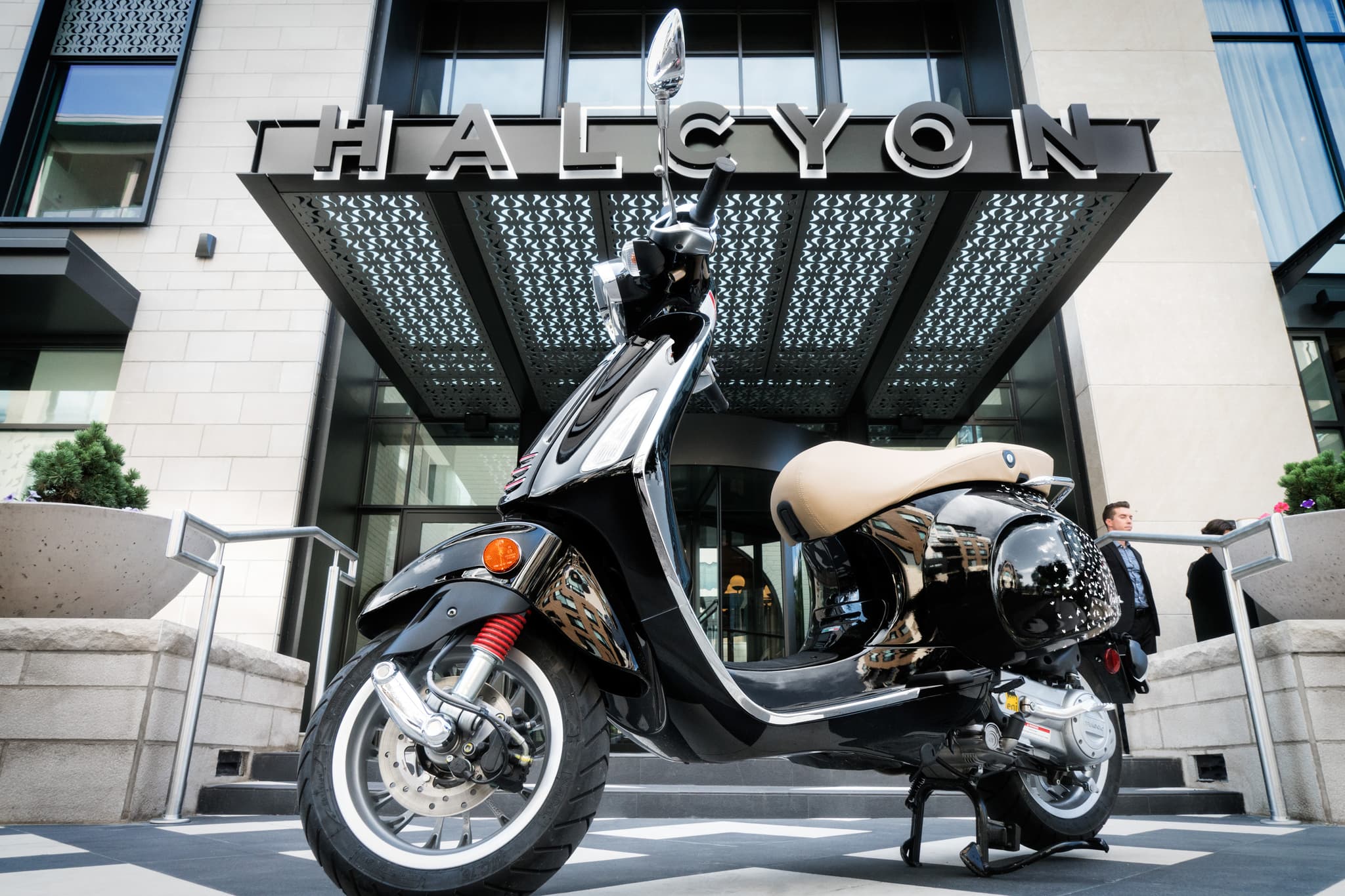A photo of a shiny black moped with a tan seat is parked on the checkered sidewalk in front of the Halcyon's large awning sign on a sunny day.