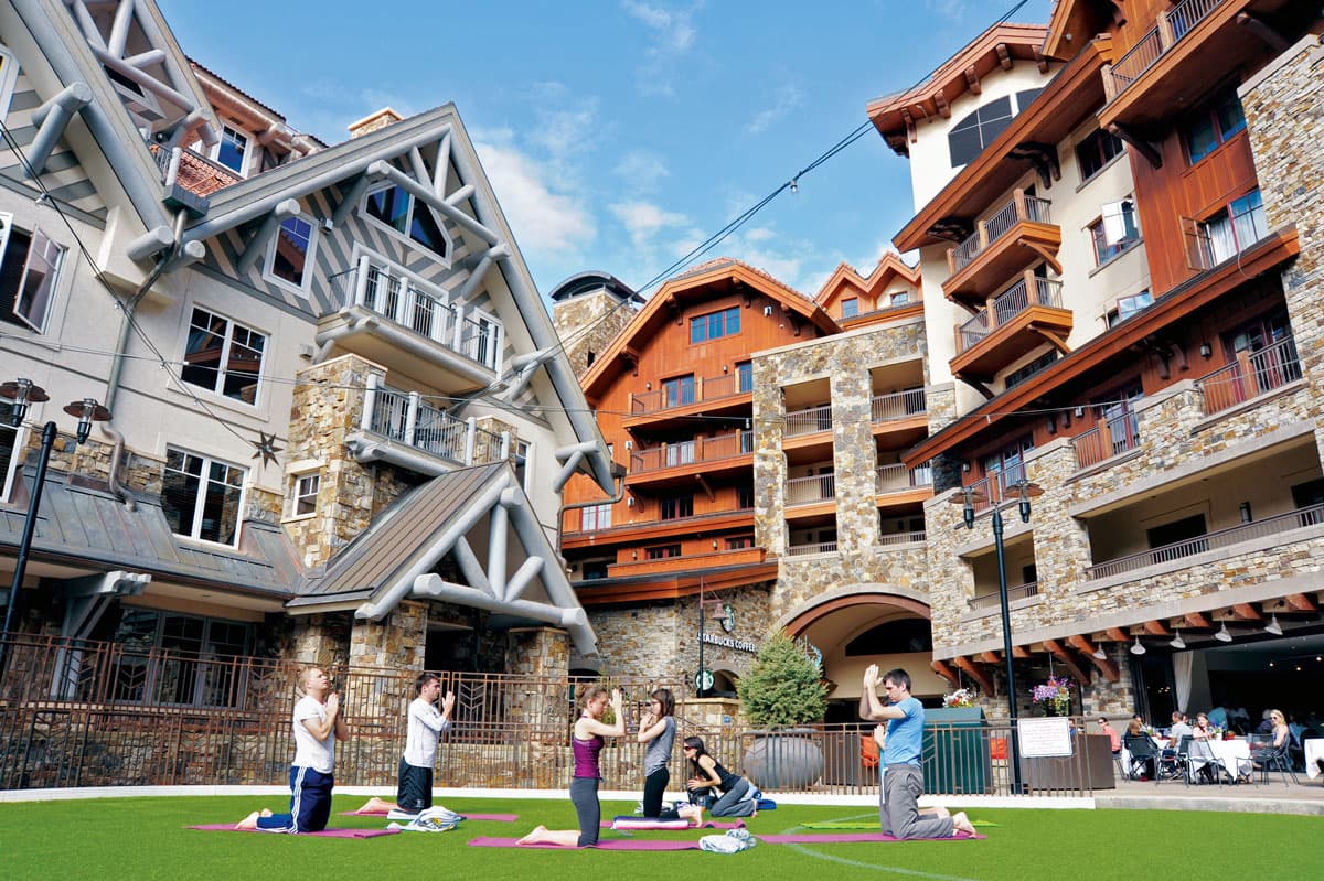 A small group of people on pink yoga mats do a yoga post on a bright-green lawn. Behind them is a variety of European-style multi-storied lodges adorned in stone and painted wood. There are people eating at restaurants around them. The sky is blue and has wispy white clouds.