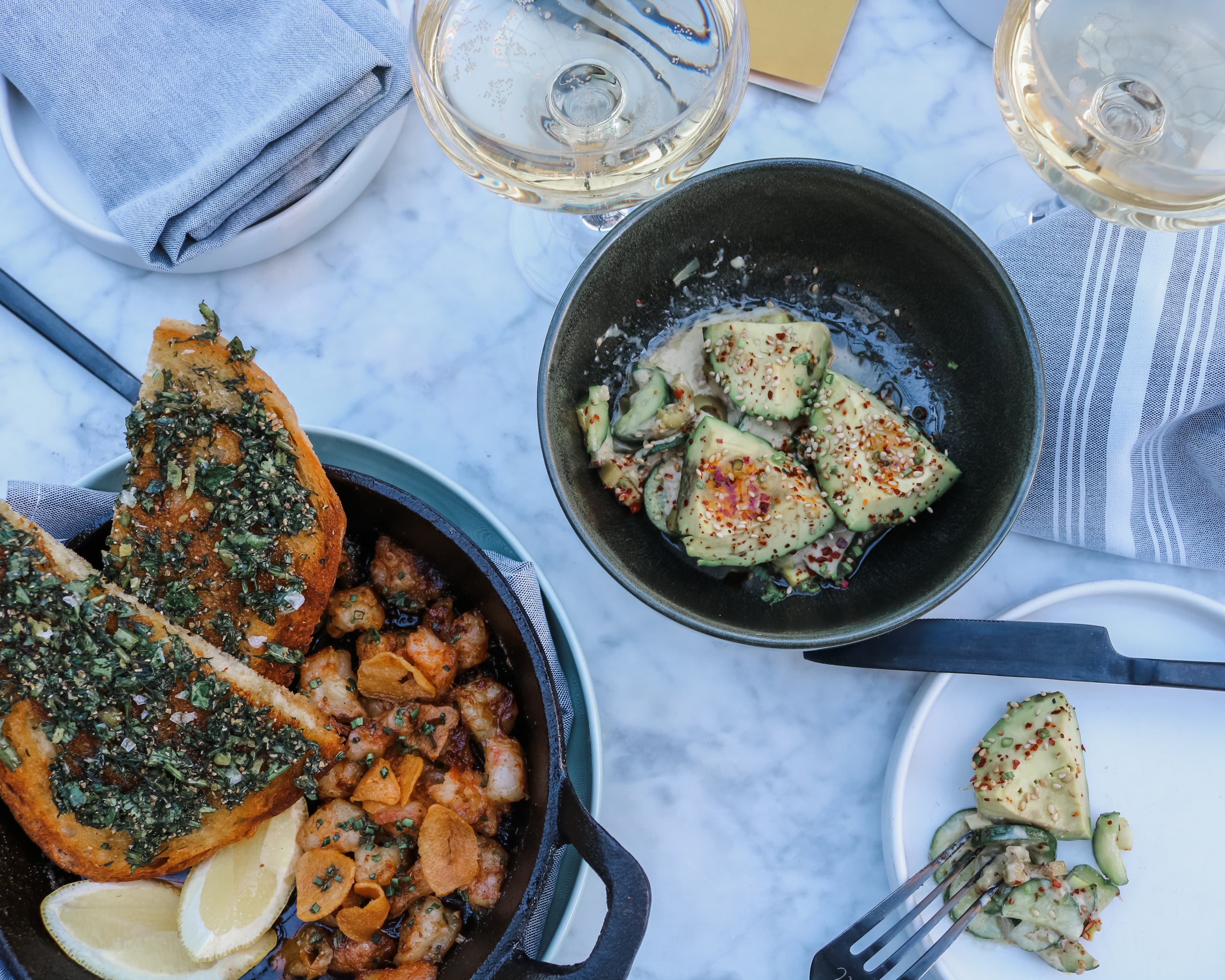 A straight up photo of two dark bowls full of colorful food, including avocados sprinkled in sesame seeds and chili flakes, slices of toast covered in chopped greens and what looks like roasted shrimp and small potatoes. There are glasses of white wine on the nicely set table as well.