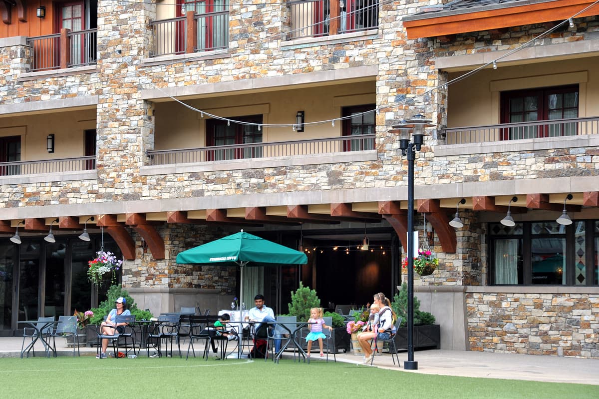 A few families with children are sitting on lawn chairs under a green umbrella outside of a dimly lit restaurant. Above them is the stone-adorned tall multi-storied building of the Madeline Hotel & Residences in Telluride, Colorado.