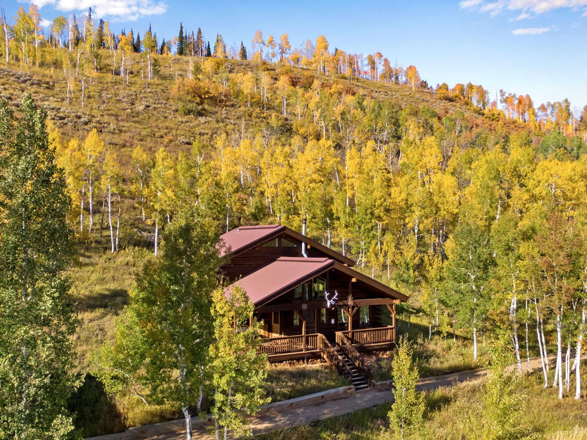 A grassy hill takes up most of the photo, covered in green-and-yellow aspen trees under a dusty blue sky. In the middle of the hill is a log cabin with maroon roofs and a staircase leading to the dirt path in front of it, going perpendicular.