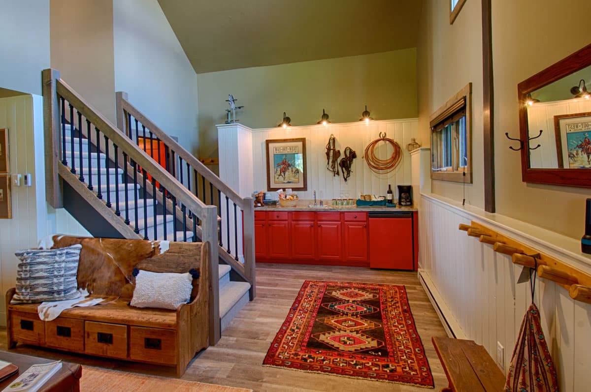 A warm-colored, large room with a small kitchen in the back of the photo. The cabinets in the kitchen are red and above the sink hangs a framed photo, horse-riding equipment and a rope for decoration. There are stairs leading up to another level. In front of the stairs is a wooden bench decorated with two throw pillows. The room is lit with natural light as well as light from the fixtures in the kitchen.