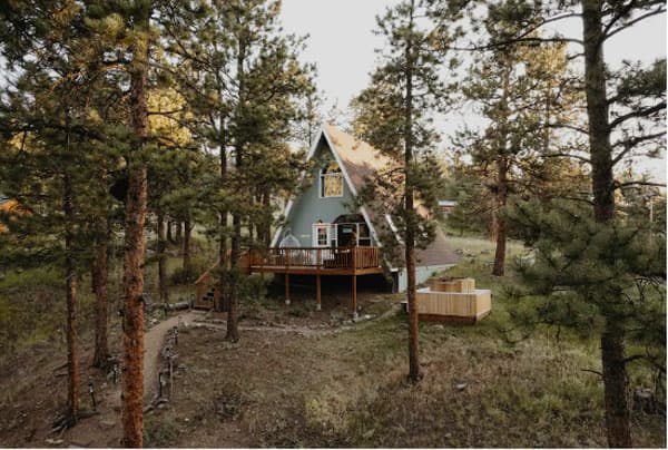 A clearing in a pine tree forest with a short winding path leading to a house with an a-frame build. In front of the white front door is a dark wood patio. Beside the house on the ground is a large cedar hot tub. The sun is shining on the side of the house.