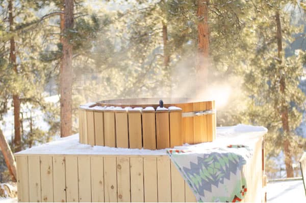 A two-tiered light brown cedar hot tub steams with the hot water inside. There is a patterned towel lying on the lower level of the hot tub. There is a light layer of snow on the first layer. In the background is sunlight filtering in through green-and-brown pine trees.