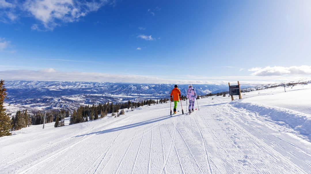 An adaptive skier and another skier pause on a ski slope to view the blue sky and mountains beyond