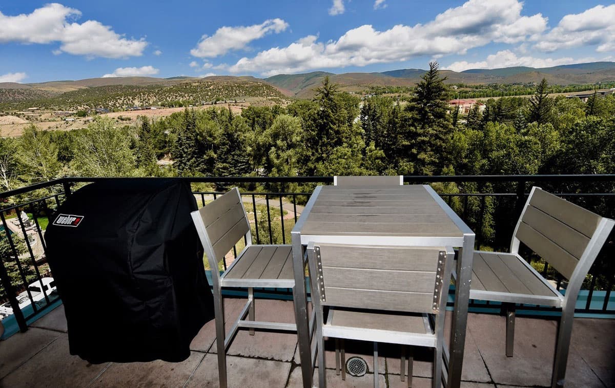 A photo of the balcony at the two bedroom river view condo at The Inn at Riverwalk. There's a covered grill next to the four-chaired table. The view in the distance is that of pine trees and distant rolling mountains. The sky is partly cloudy but the sun is shining.