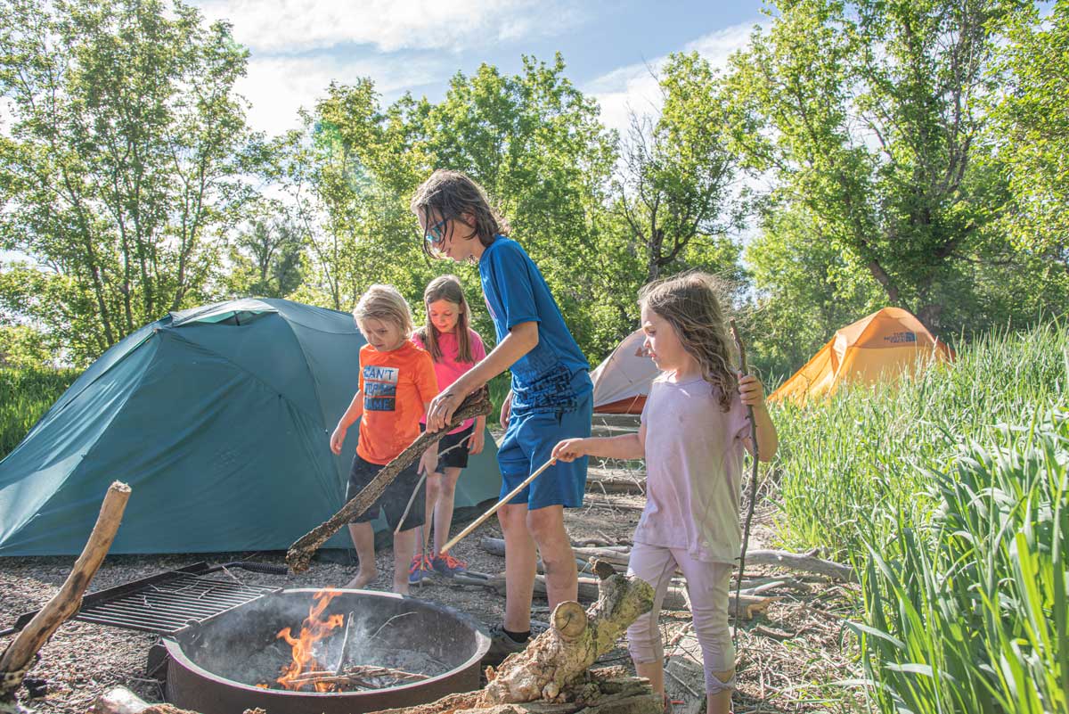 Four children tend to a campfire by a tent with green-leafed trees in the background at Yampa River State Park.