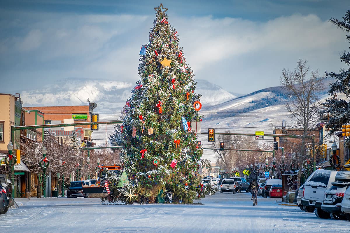 A very tall Christmas tree decorated with ribbons in the middle of a snowy, small-town street