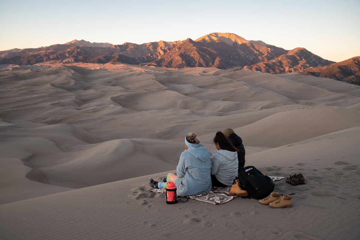 Two people sit closely on the dunes of Great Sand Dunes National Park and Preserve and look at the bordering Sangre de Cristo Mountains in Alamosa Colorado
