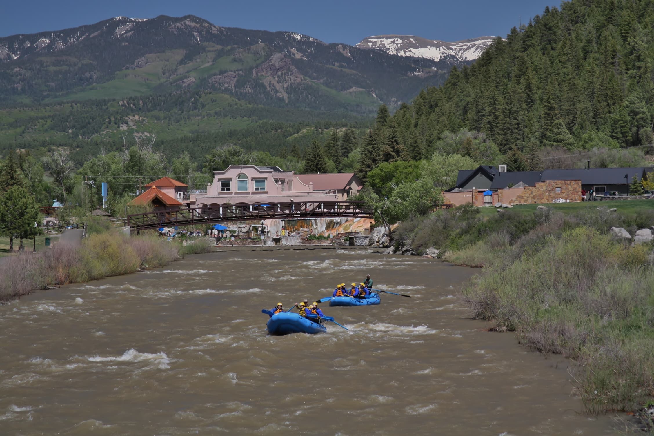 A blue raft filled with people floats down a river in Pagosa Springs with mountains in the background