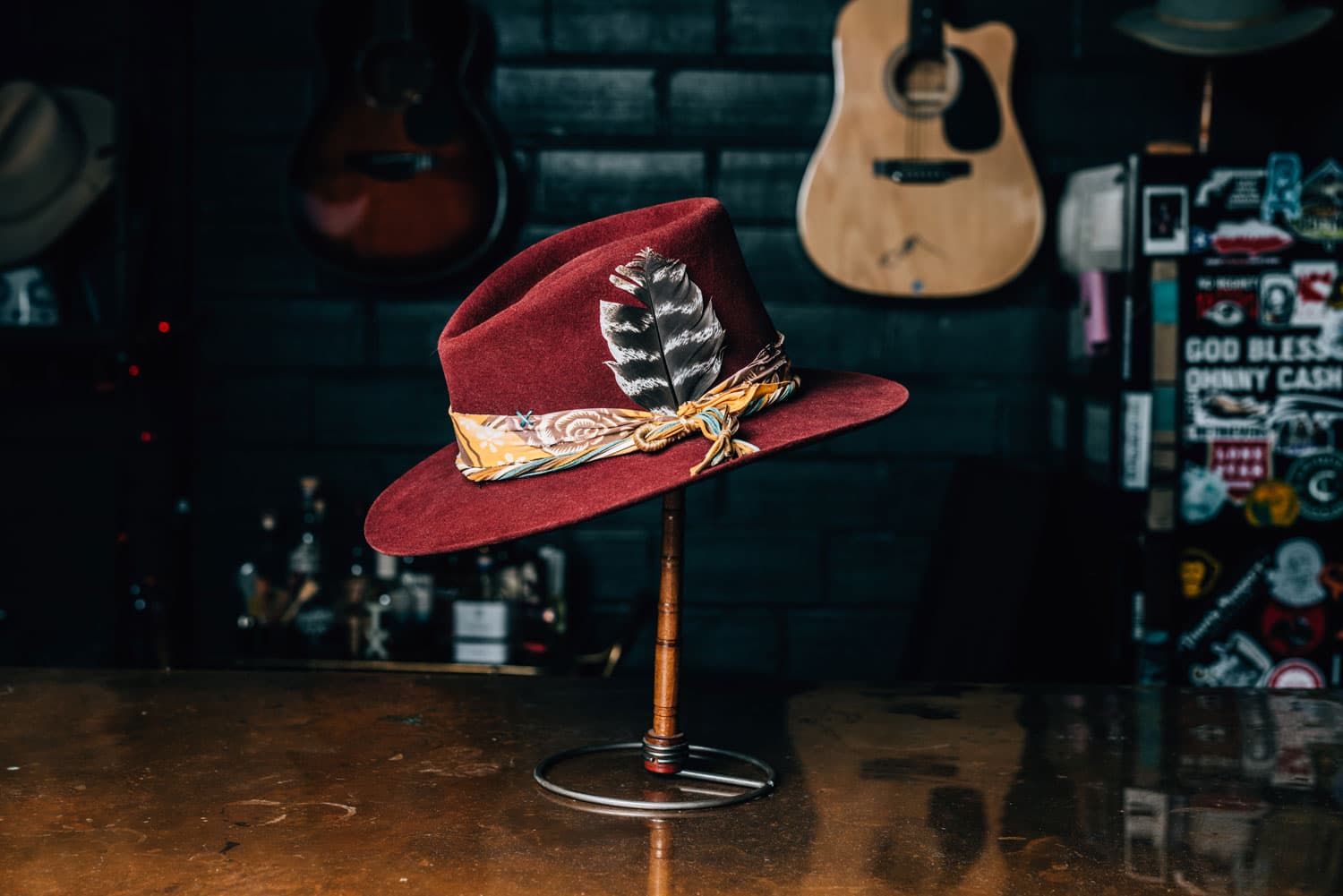 A wide-brimmed hat featuring a printed cloth and striped feather around the head piece is made of maroon velvet and sits on a hat stand at Aspen Hatter in Colorado.