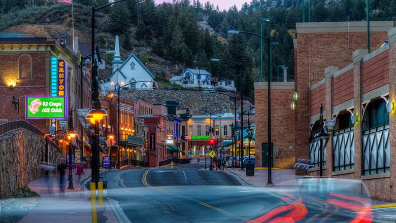 Downtown Black Hawk at sunrise. The street is empty of cars and the neon signs are ablaze. The brick buildings that sit along the winding street are all two to three stories. Up on a hill above the street sits a white church and house. There are evergreen trees above that.