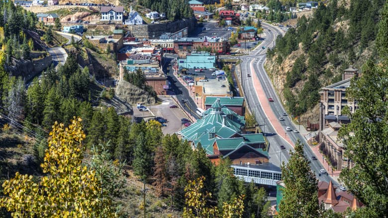 An aerial view of the gaming town of Black Hawk. We see modern green-roofed buildings and several 1800s brick buildings, as well as a two-lane highway running through town