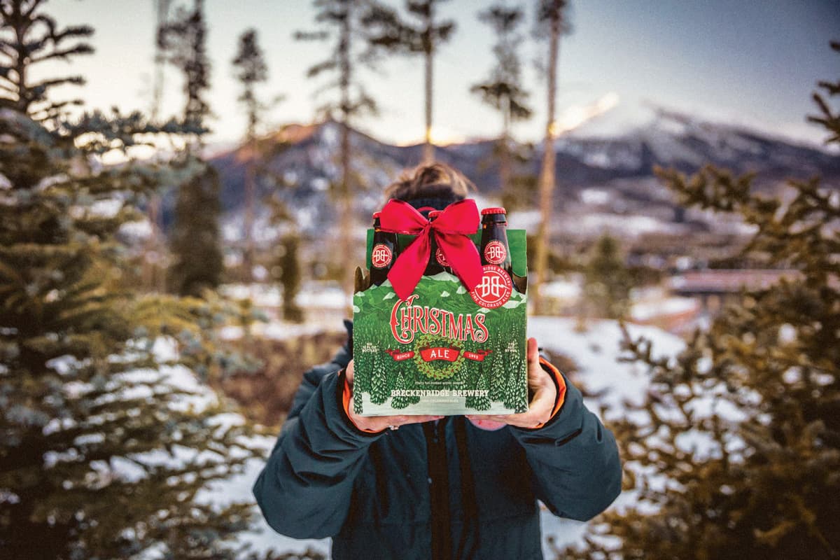 A person in a deep-green winter coat holds up a six-pack of holiday beer from Breckenridge Brewing in Littleton, Colorado. They are surrounded by snow and pine trees.