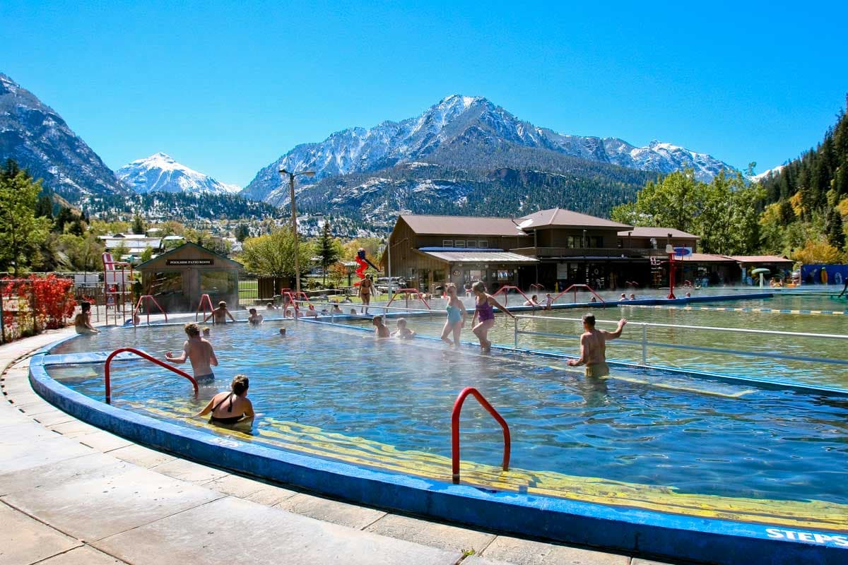 People sit and wade through the steaming water at Ouray Hot Springs. In the distance snow-covered mountains sit beneath a bright-blue sky. 