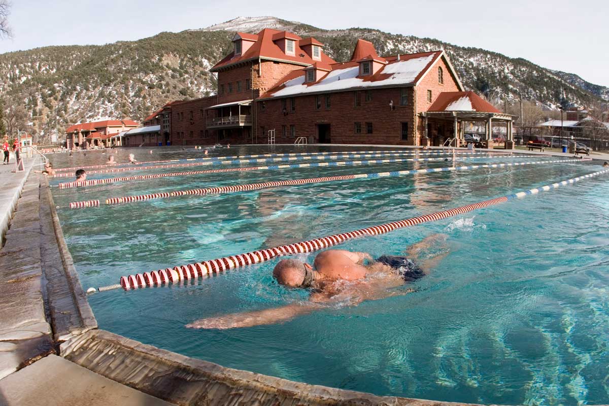 A person swims through the teal water at Glenwood Hot Springs. The historic red-brick building with a red-roof sits under a tree-covered mountain in the background.