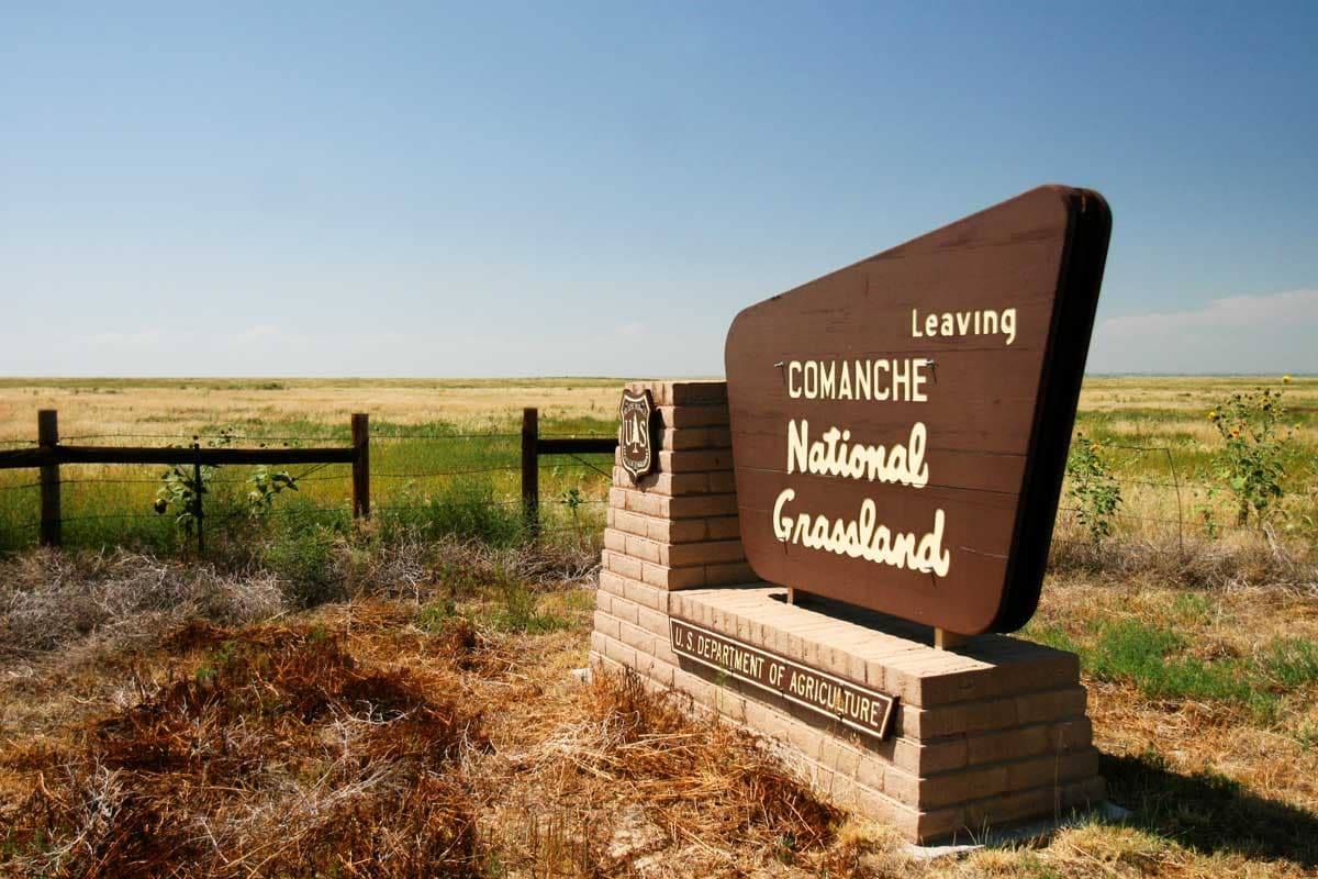 A blue sky with a haze that has a green field with a wooden fence sits behind a "Comanche National Grassland" brown sign.