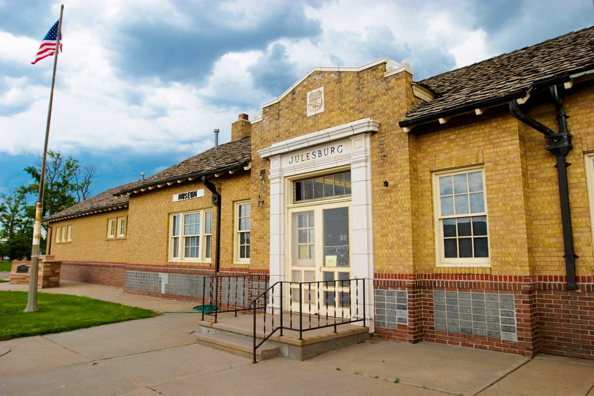 The yellow brick Julesburg Depot Museum sits under a cloudy blue sky with green grass and a flagpole with an American flag blowing in the wind out front.