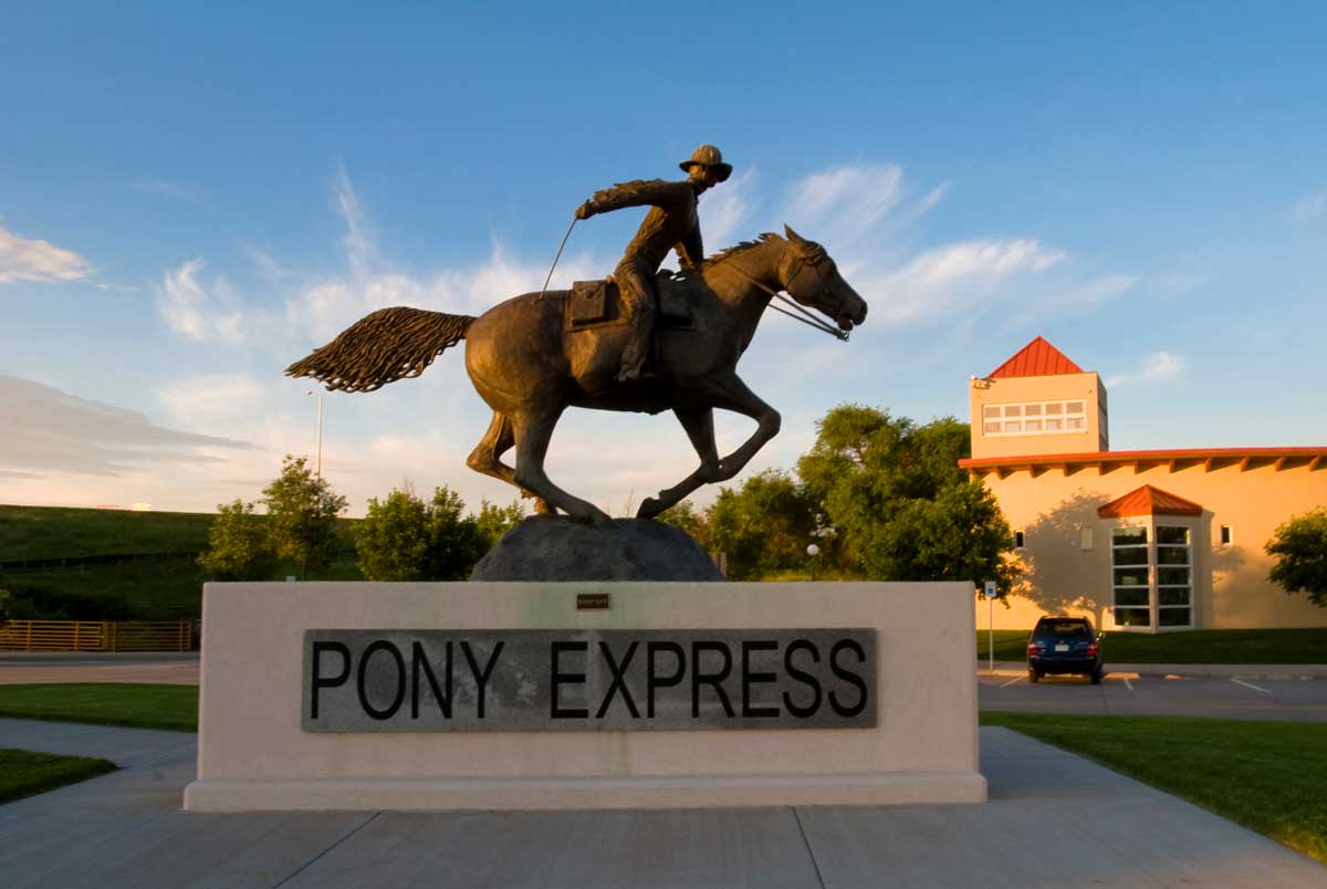 A monument that has a person riding a horse sits atop a base that says "Pony Express" sits under a blue sky at golden hour.