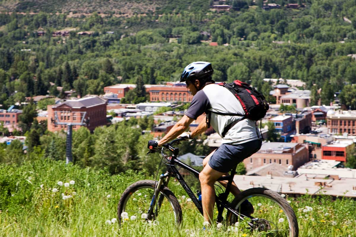 A person mountain bikes through tall green grasses and wildflowers above downtown Aspen.