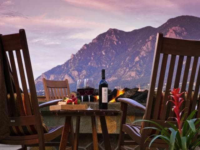 Two outdoor rocking chairs face a mountain at sunset with a bottle of wine on the table in between